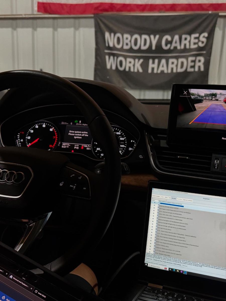 Audi Q5 interior with the dash cluster active and a "Nobody Cares Work Harder" banner in the background, ODIS open to the control modules test plan on a connected laptop